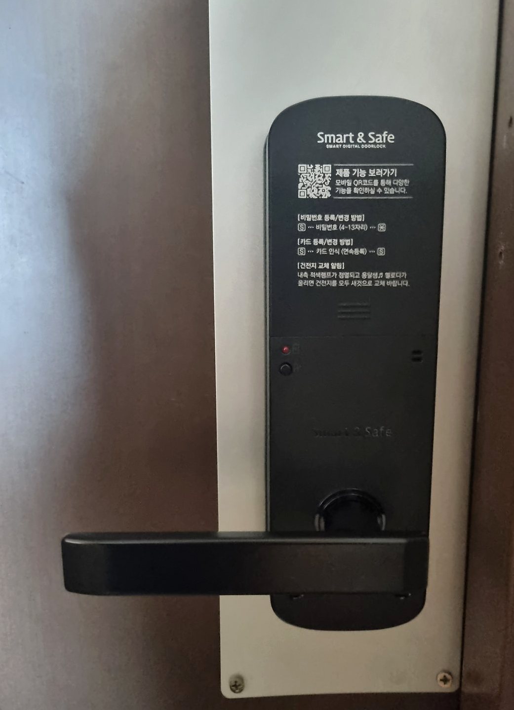 남동구 논현1동 출장 — 갈색 문의 흰 받침판 위에 붙은 검은 도어락 본체와 붉은 불빛, 왼쪽으로 뻗은 막대형 손잡이